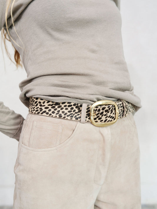 A close-up of a person wearing a leopard print belt with a large gold-tone buckle, styled with a taupe long-sleeve top and light beige pants.