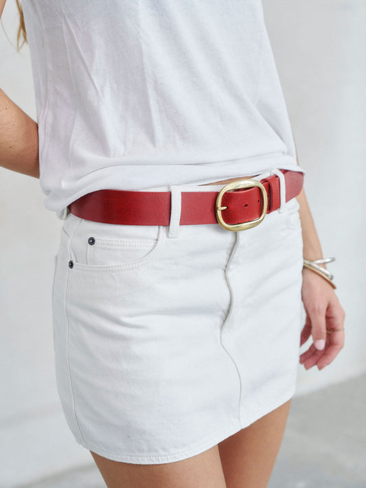 A close-up of a person wearing a white t-shirt and white denim mini skirt, styled with a red leather belt featuring a gold-tone buckle.