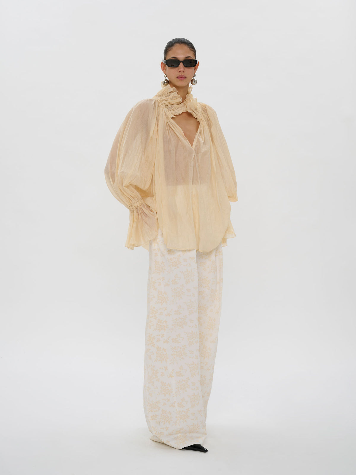 A model stands against a seamless white studio background wearing a sheer pale yellow high-neck blouse with dramatic balloon sleeves and wide-leg cream trousers with a subtle floral pattern.