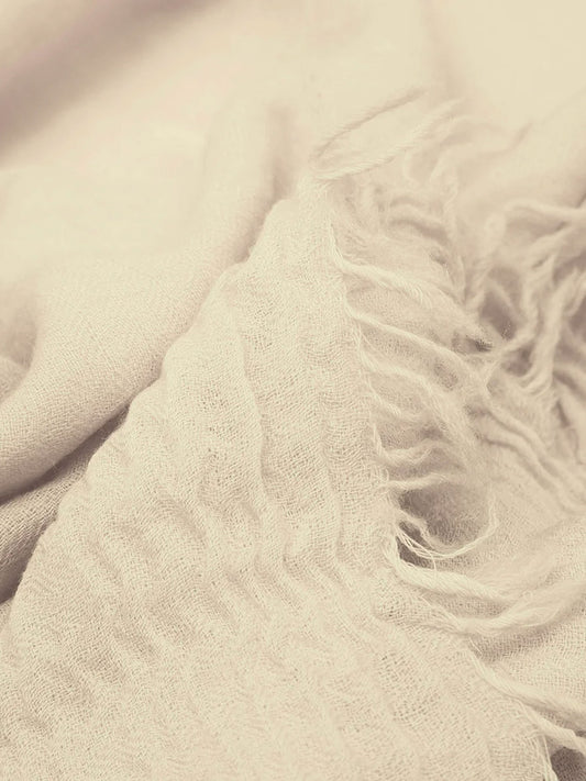 A close-up of a soft, textured cream-colored scarf with fringed edges, draped in gentle folds.