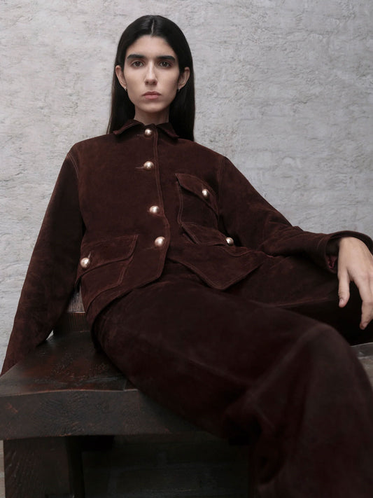 Person wearing a dark brown outfit with button details, sitting on a bench against a gray textured wall.