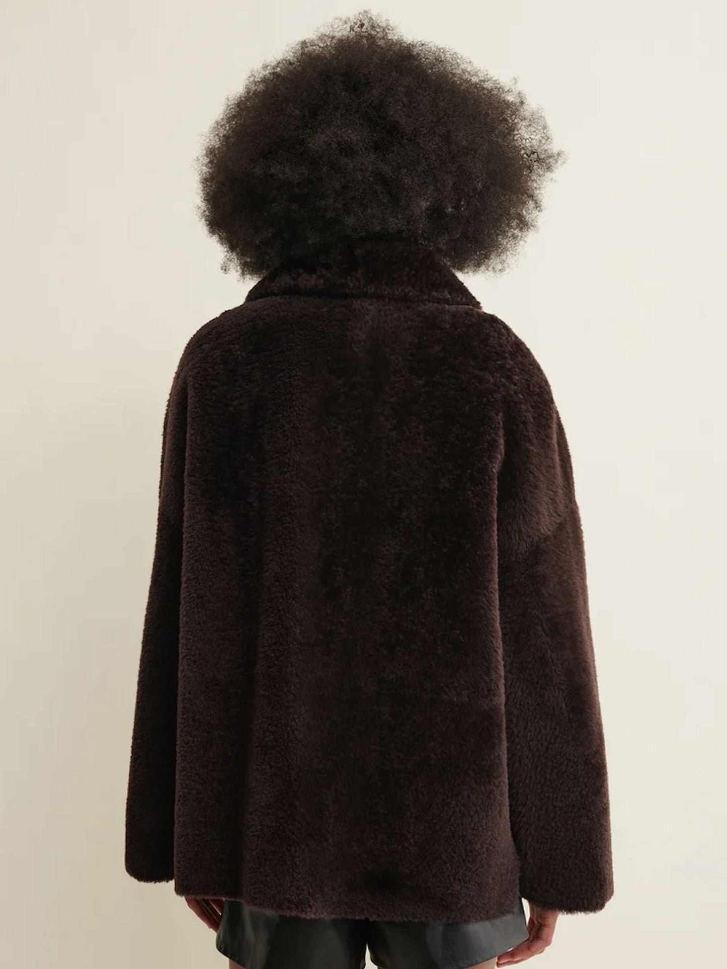 A person shown from the back wearing a dark brown fuzzy teddy-fur style jacket against a plain light beige studio background.