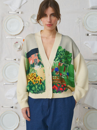 A woman wearing a cream-colored knit cardigan with a colorful, detailed landscape print featuring green trees, red flowers, and a house, paired with blue trousers. She is standing in front of a white tablecloth decorated with plates and cutlery.