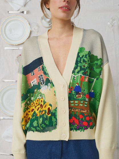 A woman wearing a cream-colored cardigan with a colorful, detailed landscape print on the front featuring a house, garden, and sunflowers, styled with blue pants against a white wall decorated with plates.
