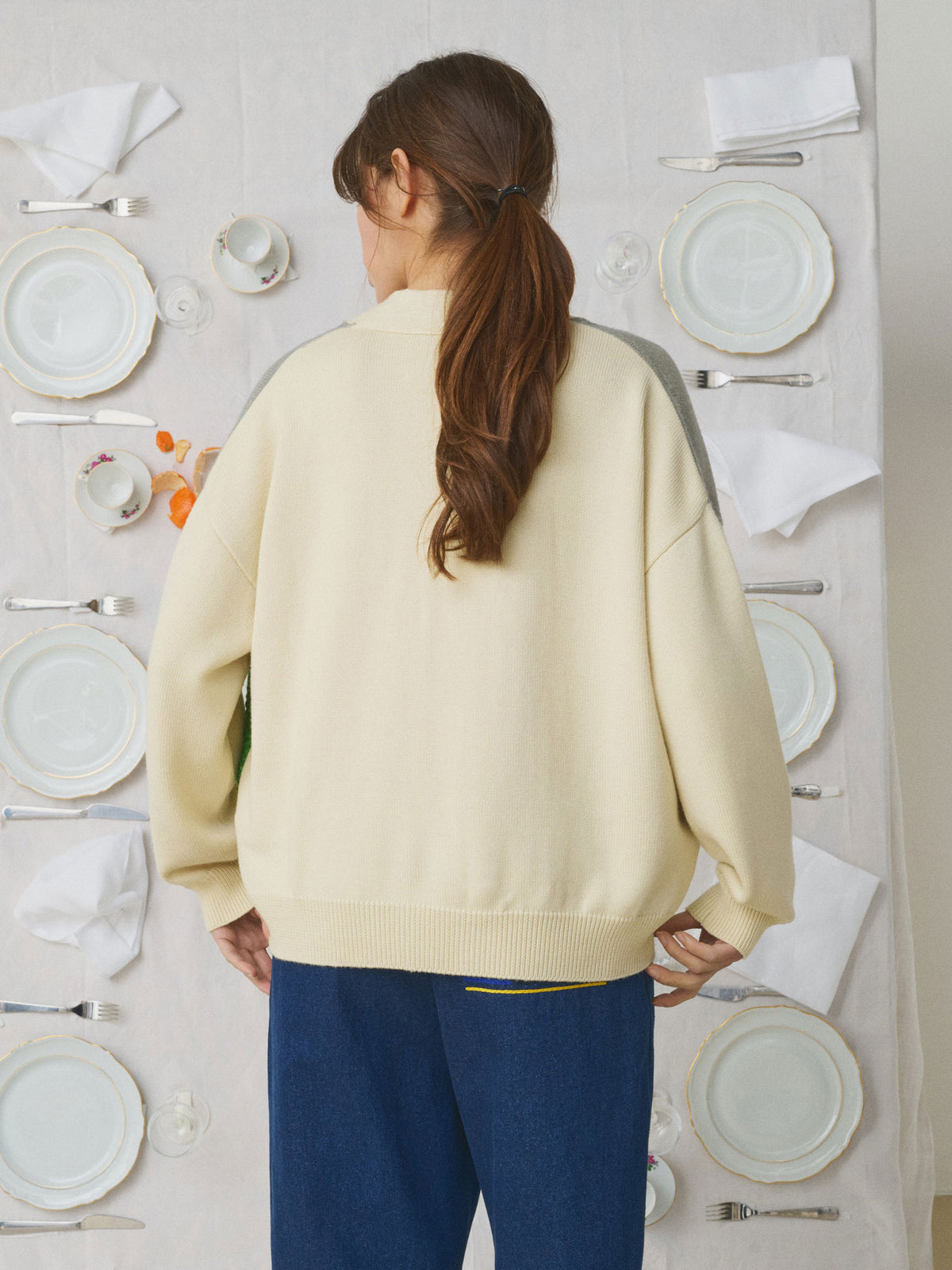 A woman viewed from behind wearing an oversized cream-colored knit sweater and blue pants, standing in front of a styled backdrop of white plates, napkins, and silver cutlery.