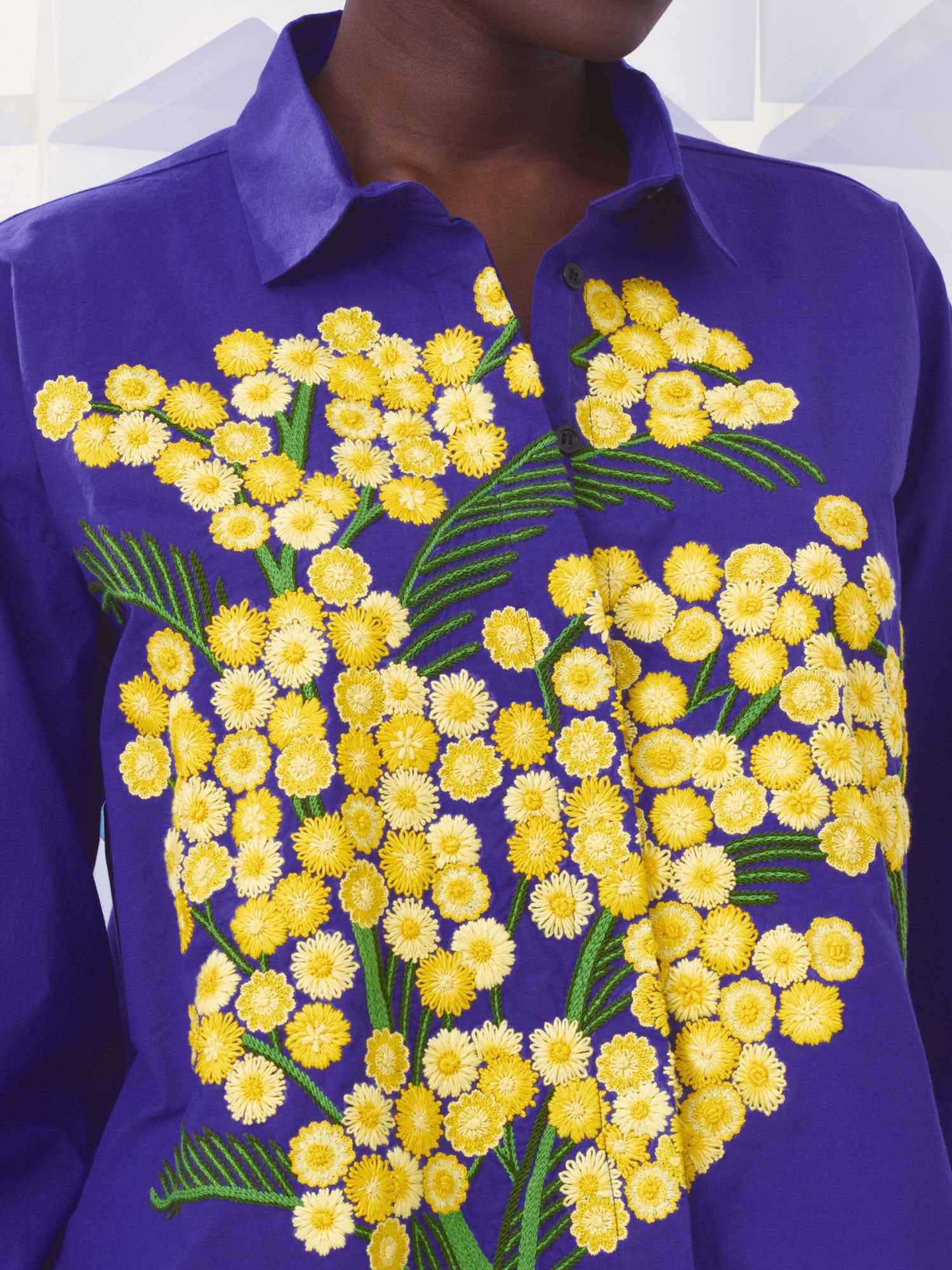 A close-up of a person wearing a vibrant blue button-up Mii shirt decorated with a large, detailed yellow and white floral embroidery pattern on the front.