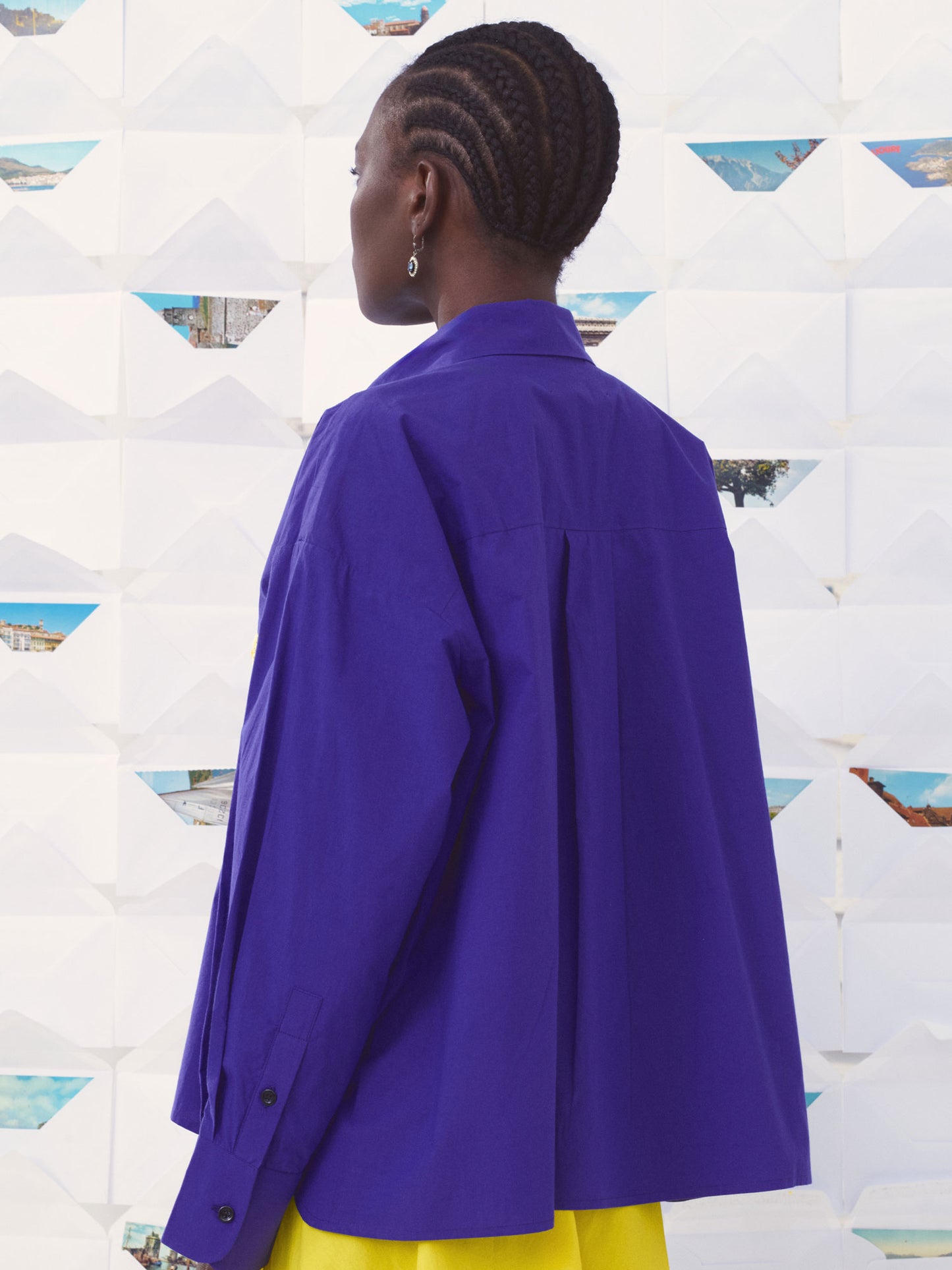 A person shown from the back wearing a bright blue oversized button-up Mii shirt and yellow bottoms, standing in front of a white geometric wall with triangular cutouts.