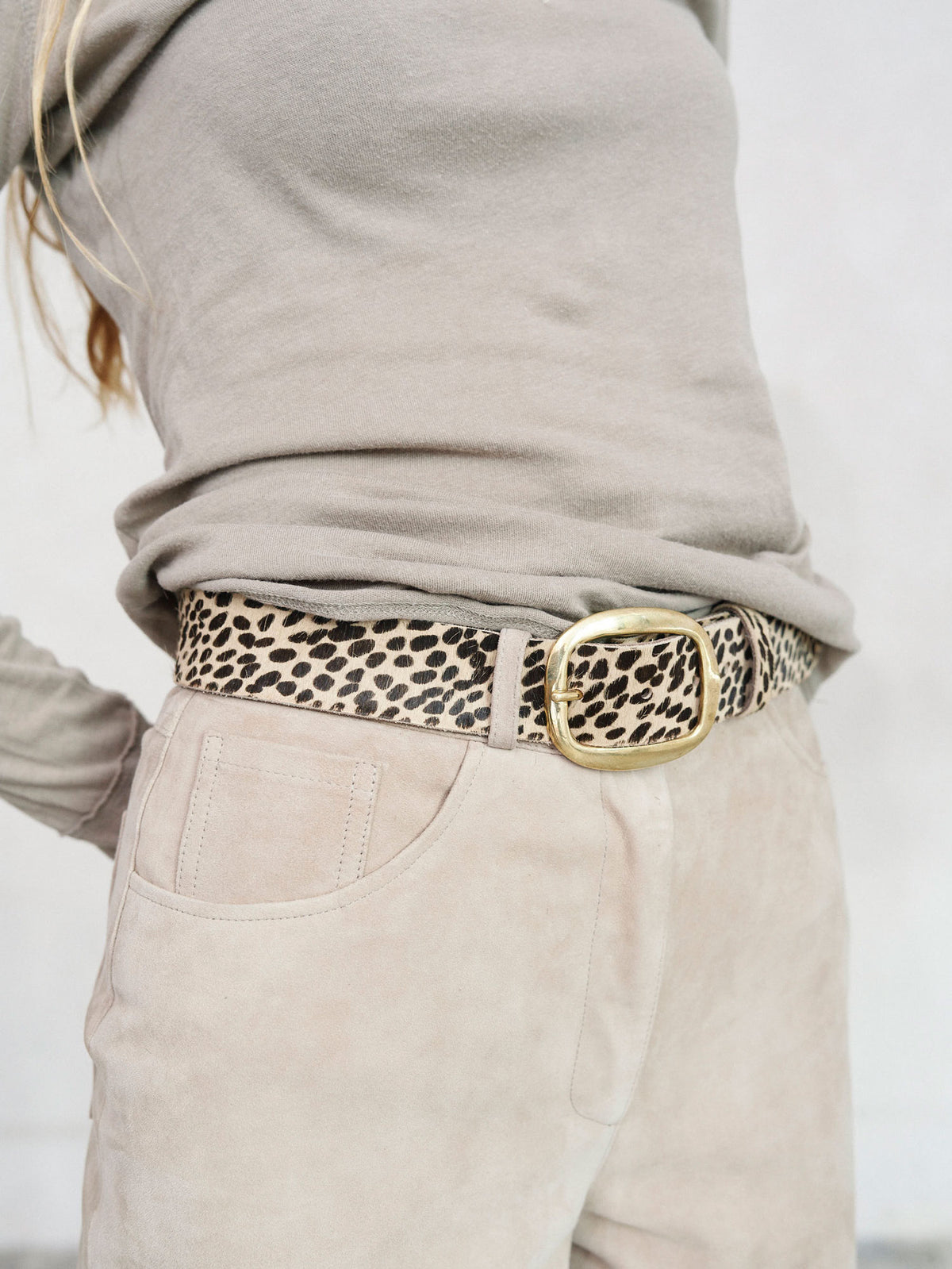 A close-up of a person wearing a leopard print belt with a large gold-tone buckle, styled with a taupe long-sleeve top and light beige pants.