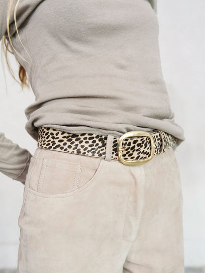 A close-up of a person wearing a leopard print belt with a large gold-tone buckle, styled with a taupe long-sleeve top and light beige pants.