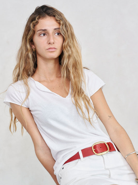 A woman with long wavy blonde hair wearing a white v-neck t-shirt and white shorts, accessorized with a red belt with a gold buckle, posed against a light gray background.