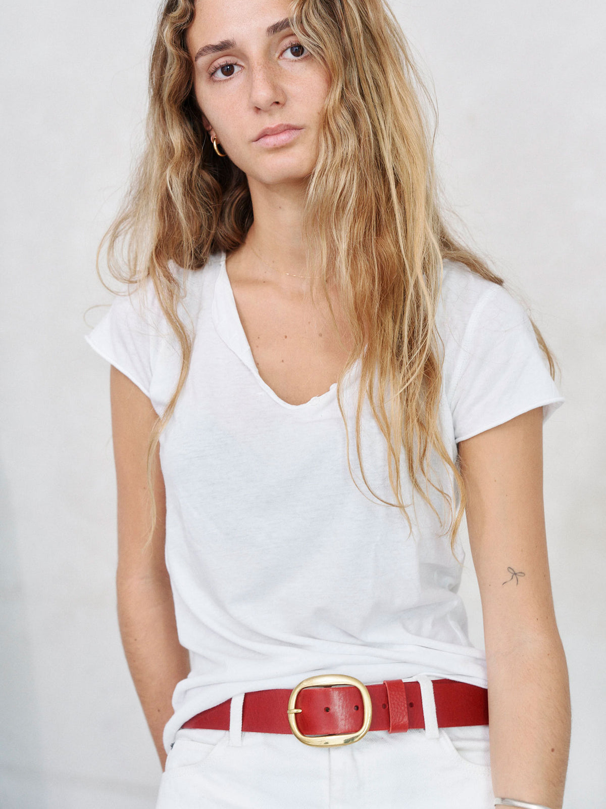 A woman with long blonde hair wearing a white V-neck t-shirt, white pants, and a red leather belt with a gold buckle, photographed against a light gray background.
