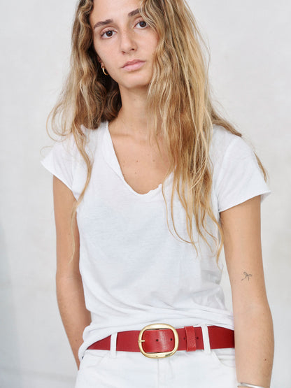 A woman with long blonde hair wearing a white V-neck t-shirt, white pants, and a red leather belt with a gold buckle, photographed against a light gray background.