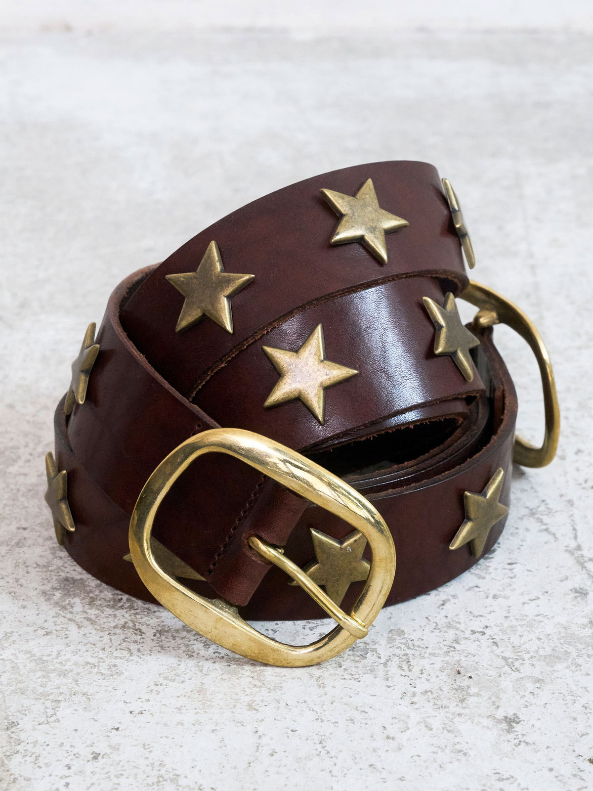A dark brown leather belt with gold-toned star-shaped metal studs and a large gold buckle, coiled on a light gray textured surface.