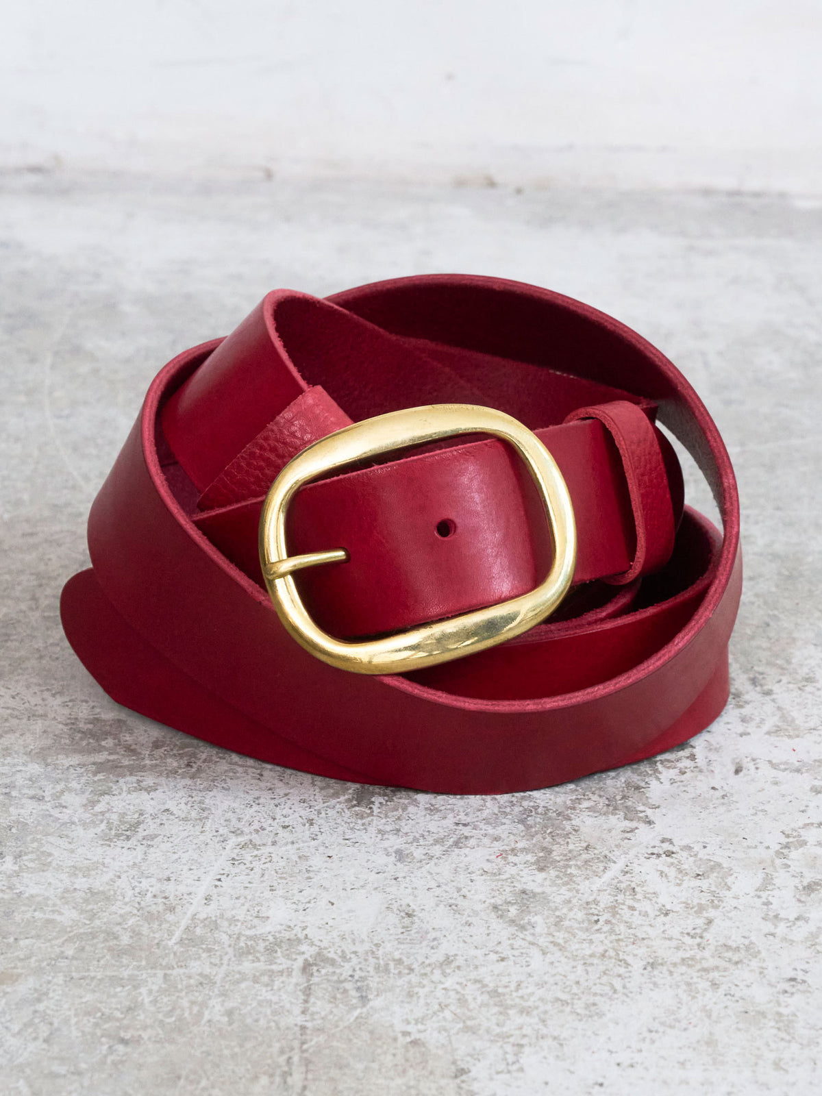 A deep red leather belt with a large, polished gold-tone metal buckle, neatly coiled on a textured light gray surface.
