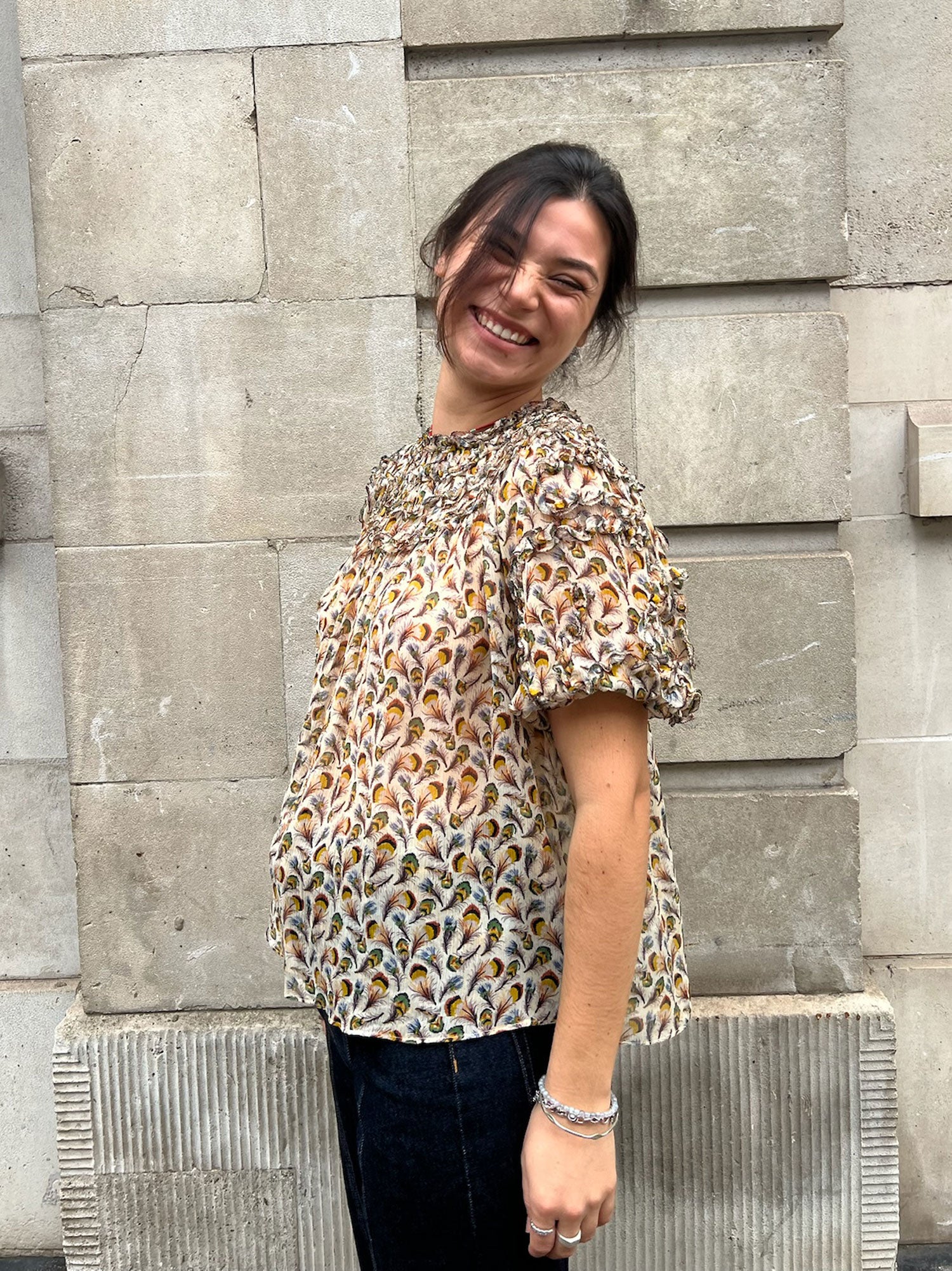 Woman wearing a patterned blouse standing against a stone wall.