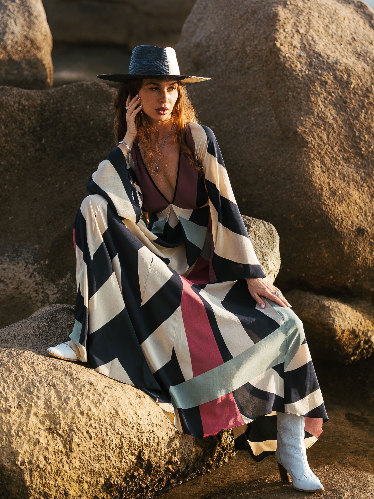 Woman sitting on rocks wearing a colorful striped dress and wide-brimmed hat.