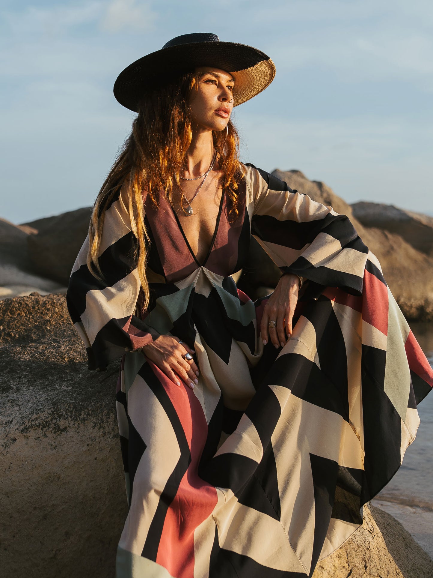 Woman in a patterned dress and wide-brimmed hat sitting on rocks with a scenic background