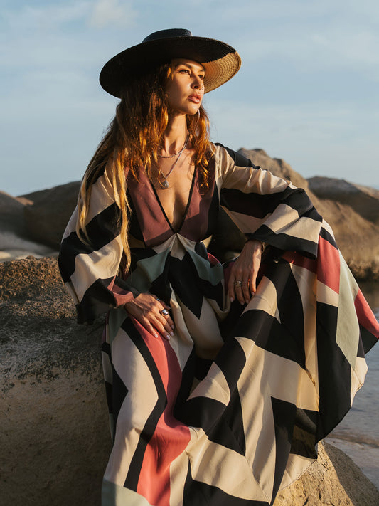 Woman in a patterned dress and wide-brimmed hat sitting on rocks with a scenic background