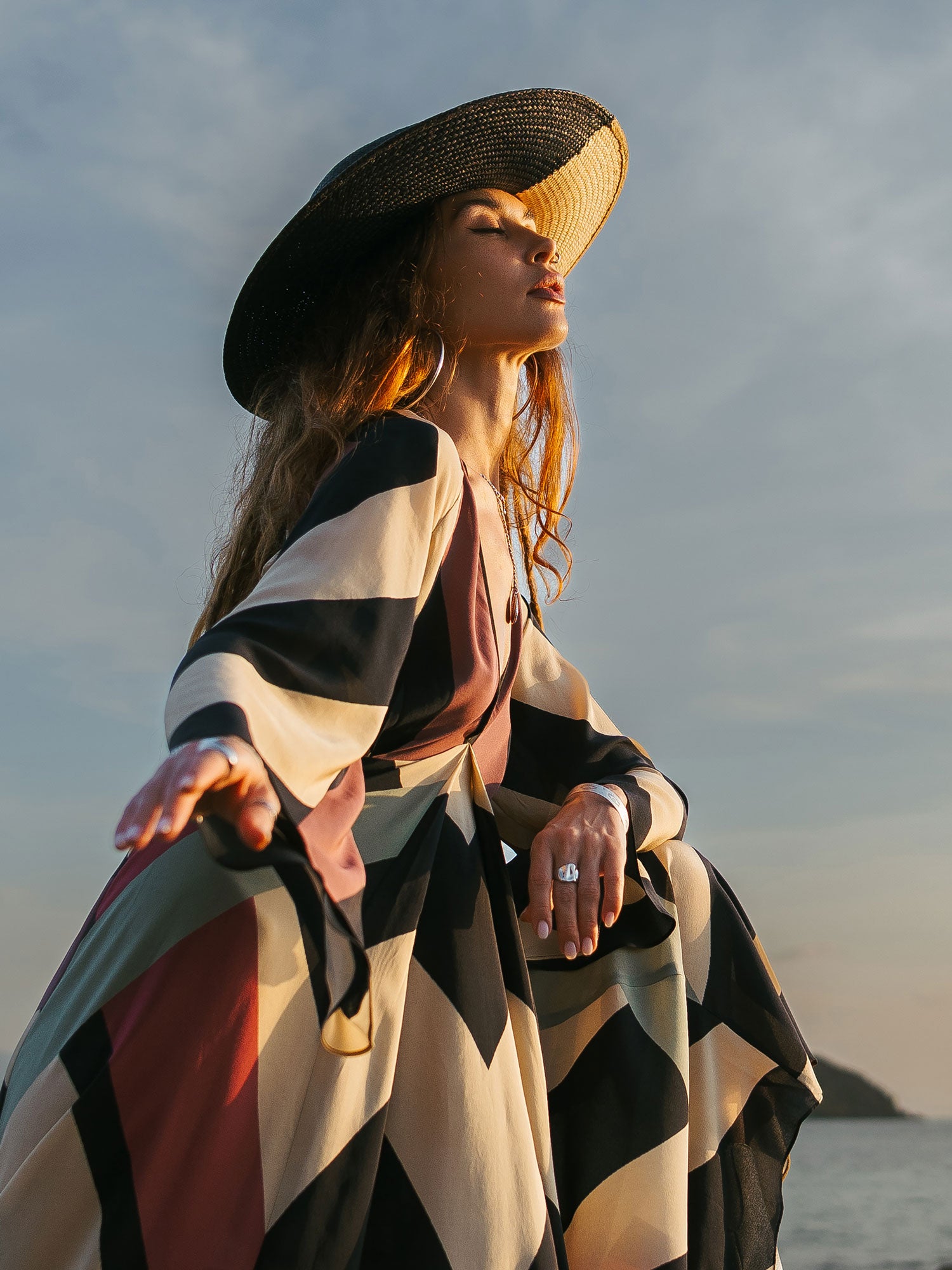 Woman wearing a striped dress and wide-brimmed hat against a cloudy sky.