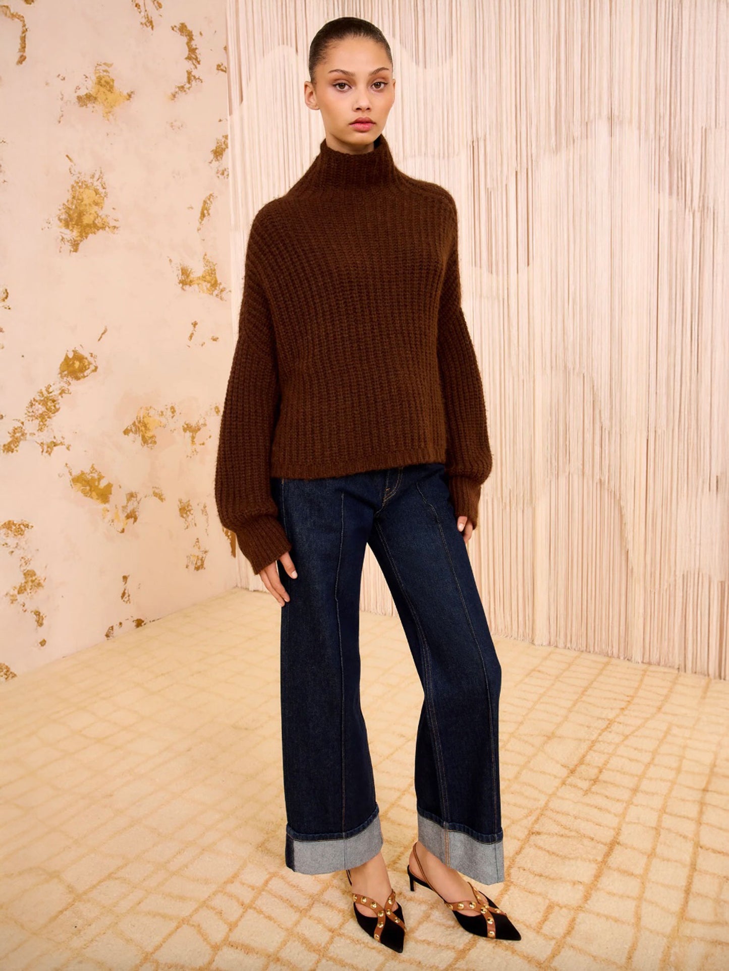 Woman wearing a brown sweater and blue jeans standing against a patterned wall.