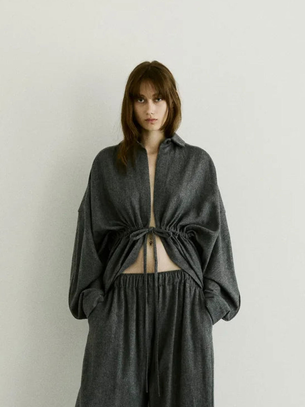 A model wearing a dark gray textured two-piece outfit consisting of a long-sleeve cropped top with a drawstring tie at the waist and matching wide-leg pants, set against a plain light gray background.