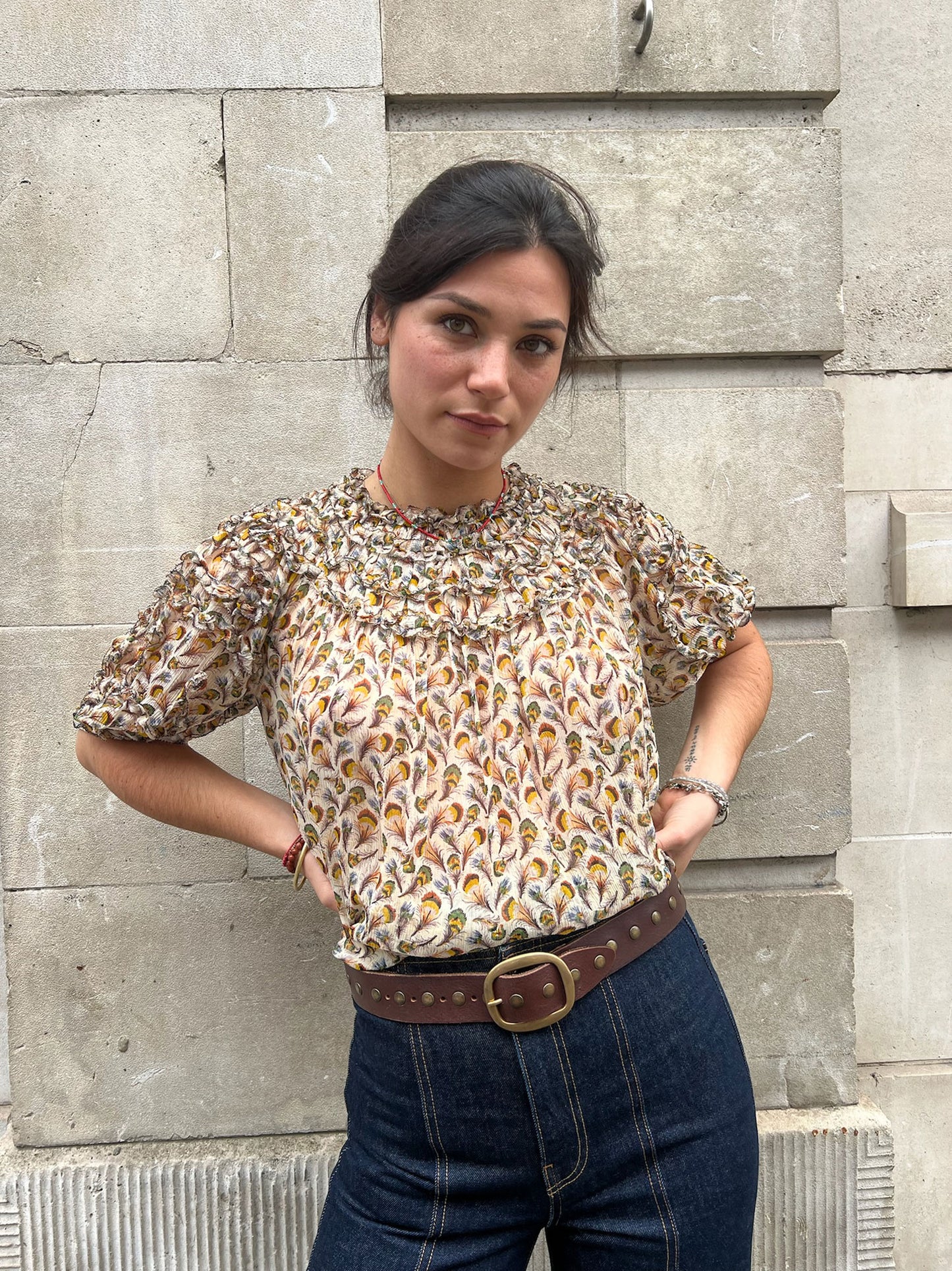 Woman wearing a patterned blouse and jeans standing against a stone wall.