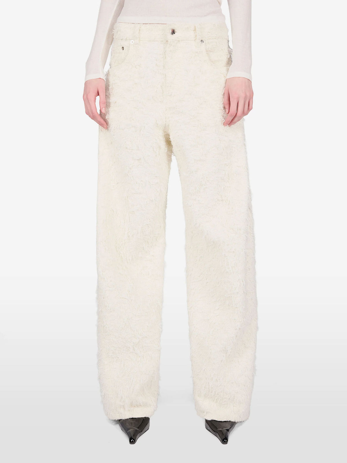 A model is shown from the waist down wearing a pair of wide-leg, high-pile textured cream pants, paired with a light beige long-sleeve top and black pointed-toe heels, against a plain white studio background.