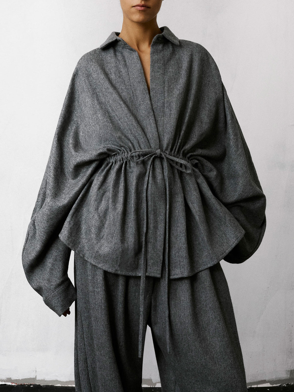 A model wearing a dark gray textured linen-blend top with a drawstring waist and matching wide-leg trousers, photographed against a light gray wall.