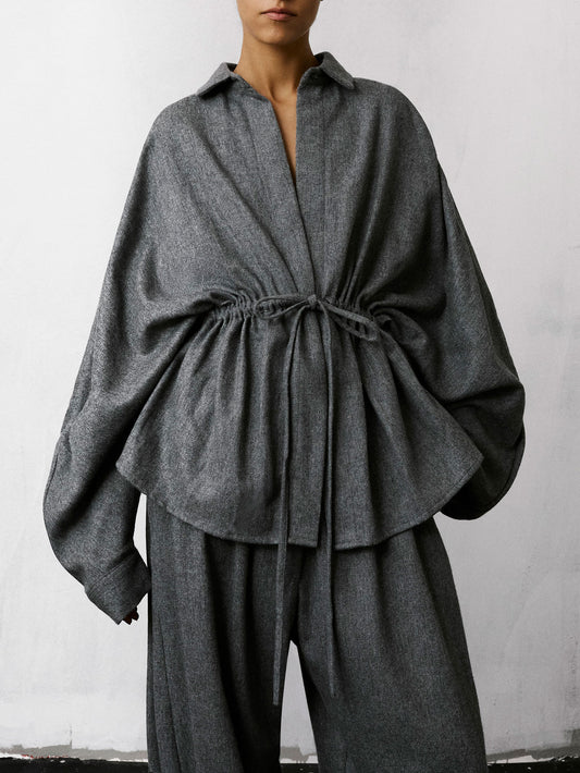 A model wearing a dark gray textured linen-blend top with a drawstring waist and matching wide-leg trousers, photographed against a light gray wall.