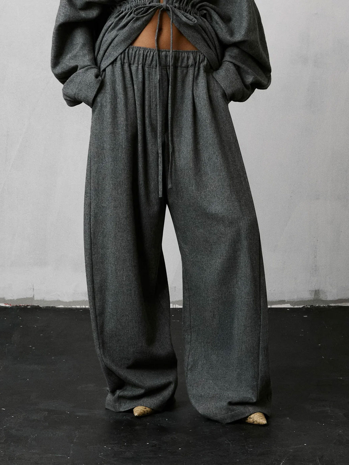A person wearing loose, wide-leg dark gray pants with a drawstring waist and a matching dark gray cropped top with a back cutout, standing against a light gray wall and dark floor.