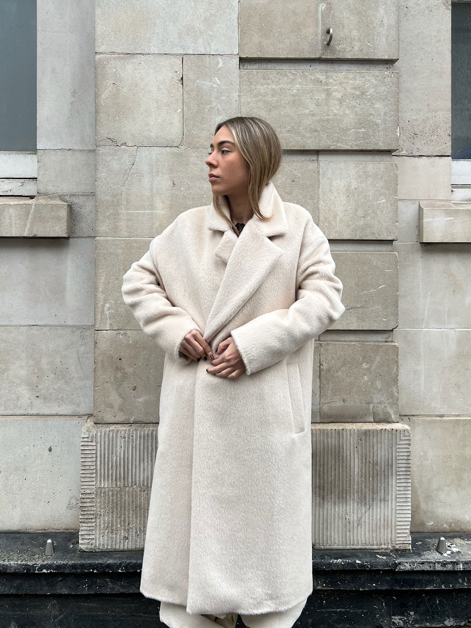 Woman wearing a long cream coat standing against a stone wall.