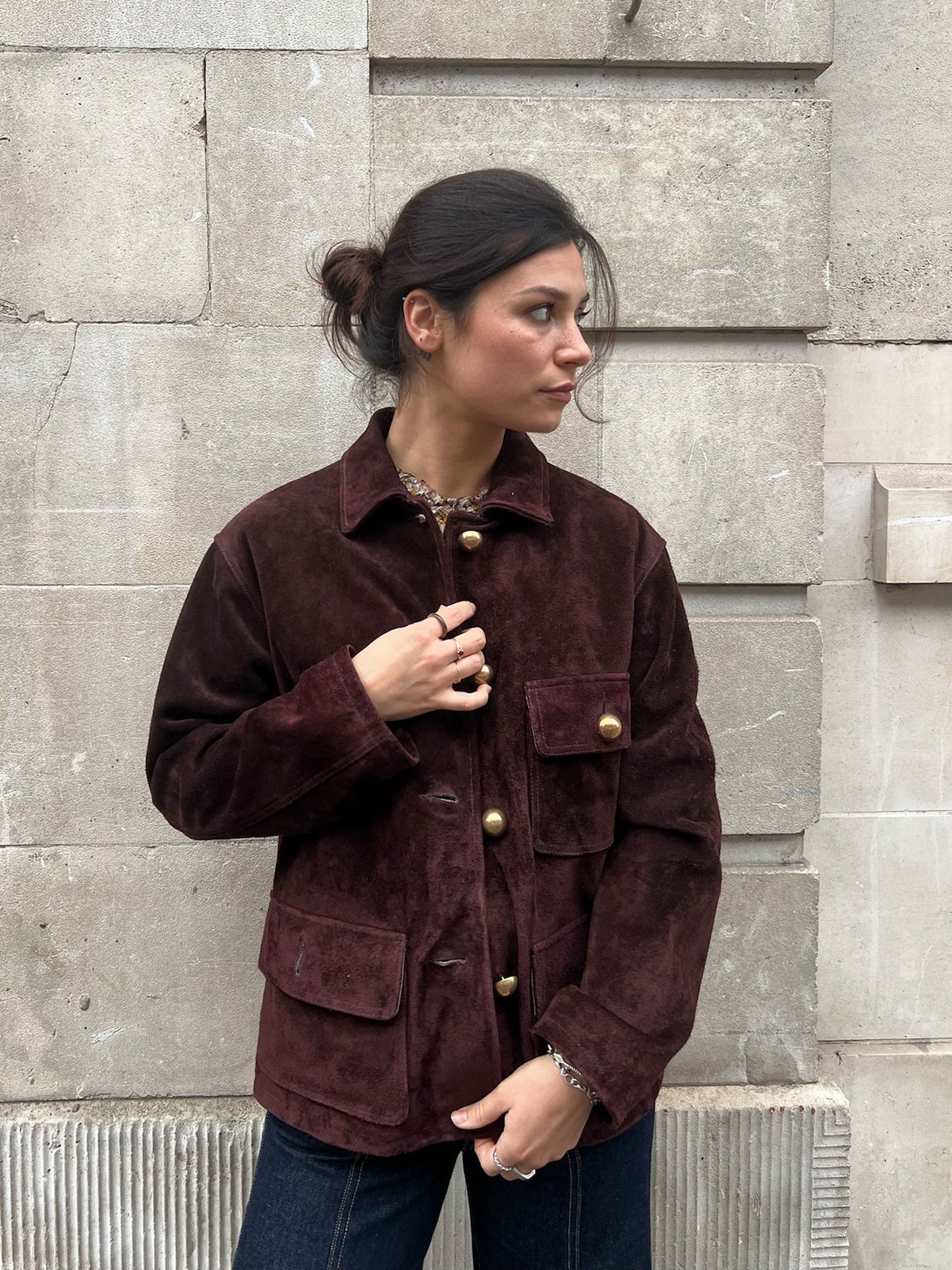 Woman wearing a brown suede jacket standing against a stone wall.