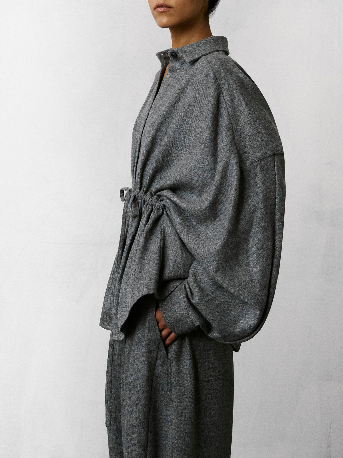 A model shown from the side wearing a dark gray textured long-sleeve shirt with a drawstring waist and matching wide-leg trousers against a light gray wall.