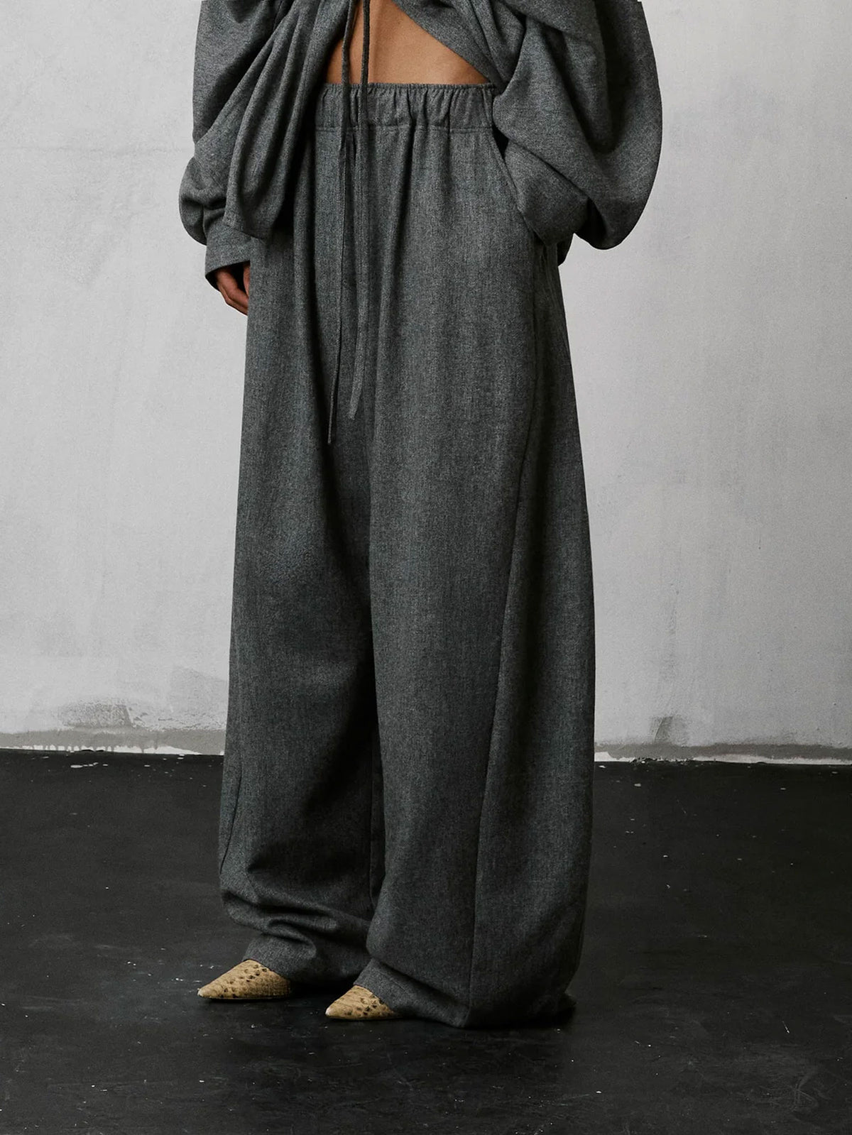 A person wearing loose, oversized dark gray lounge pants with a drawstring waist and a matching draped top, standing against a light gray wall and dark floor.