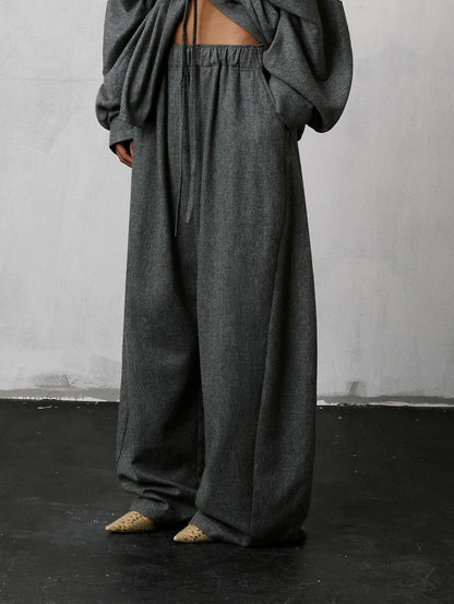A person wearing loose, oversized dark gray lounge pants with a drawstring waist and a matching draped top, standing against a light gray wall and dark floor.