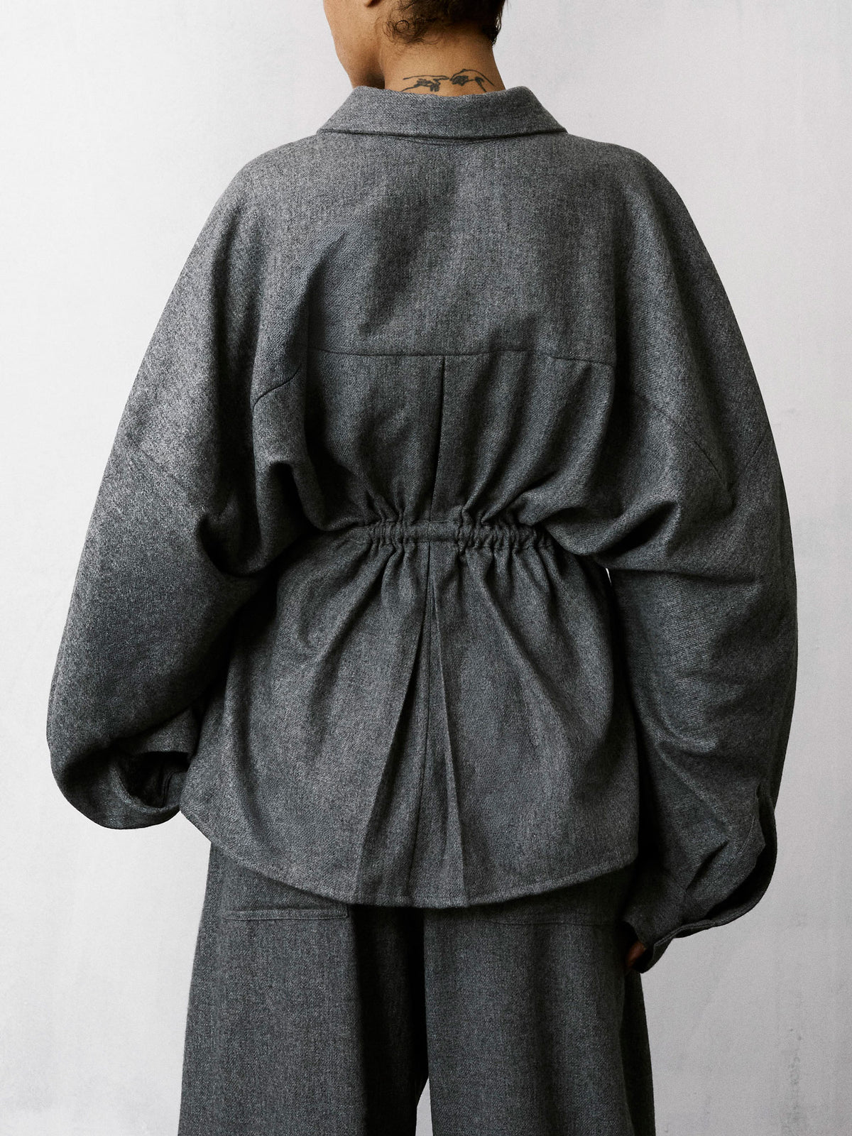 A person shown from the back wearing a dark gray oversized long-sleeve top with a gathered waist and matching dark gray trousers, against a plain light gray wall.