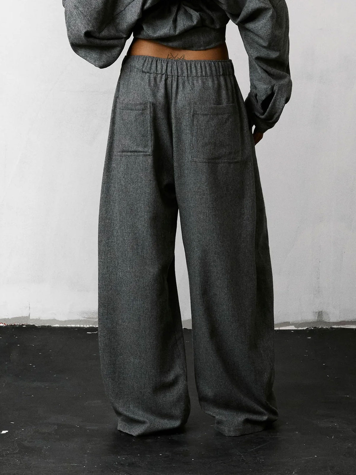 A person shown from the back wearing dark gray wide-leg trousers with an elastic waistband and large patch pockets, paired with a matching dark gray cropped top against a plain light wall and dark floor.