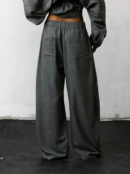 A person shown from the back wearing dark gray wide-leg trousers with an elastic waistband and large patch pockets, paired with a matching dark gray cropped top against a plain light wall and dark floor.