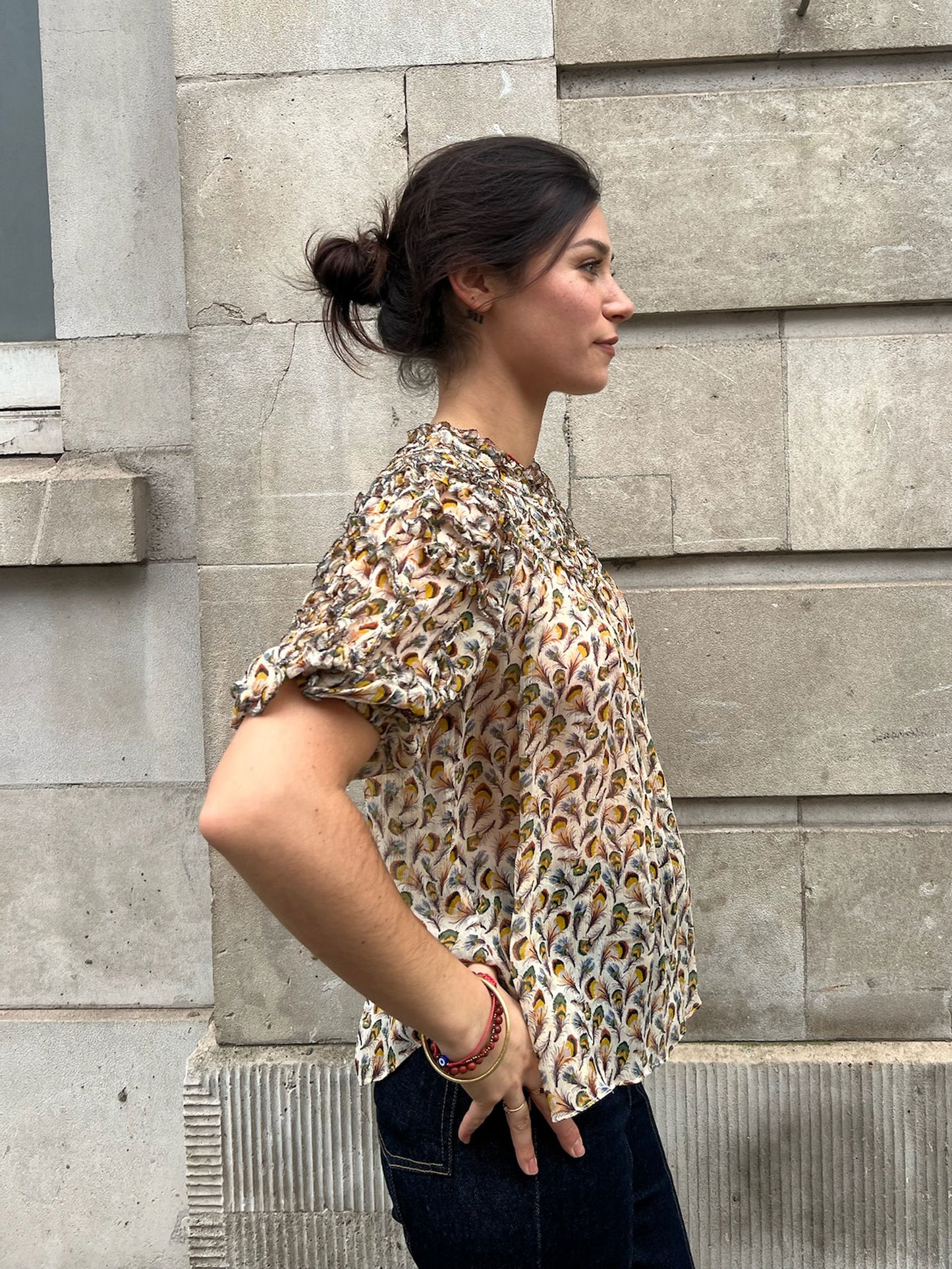 Woman wearing a patterned blouse standing against a stone wall.