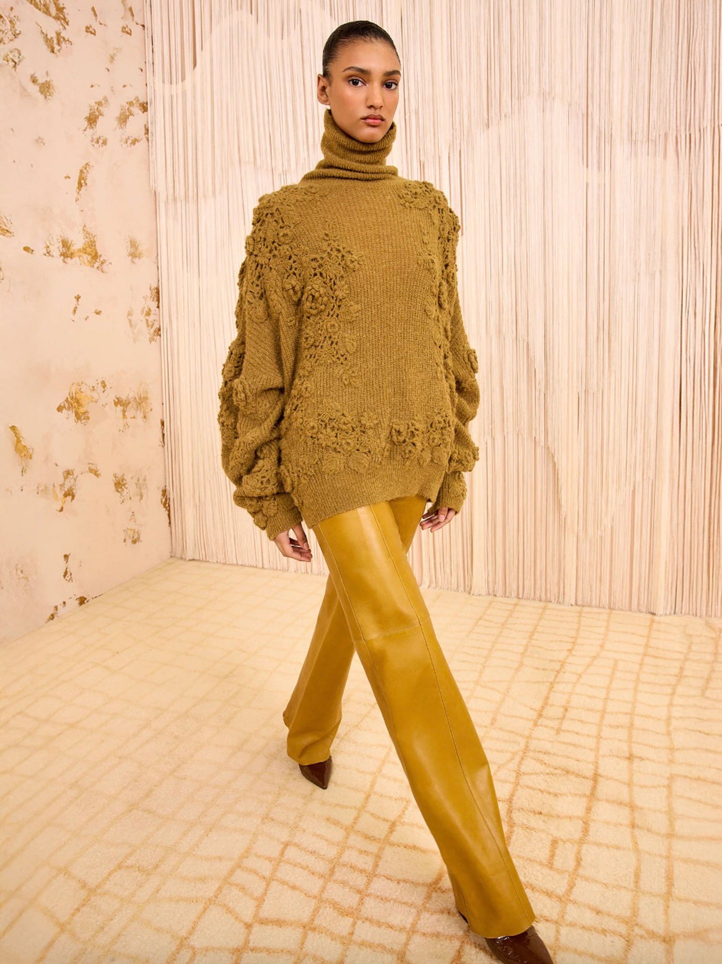 Person wearing a mustard yellow sweater and pants against a textured wall.