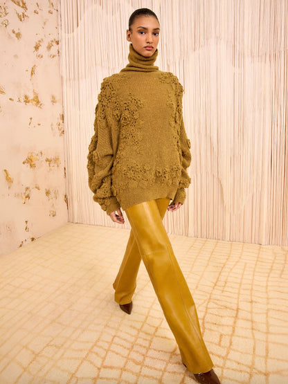 Person wearing a mustard yellow sweater and pants against a textured wall.