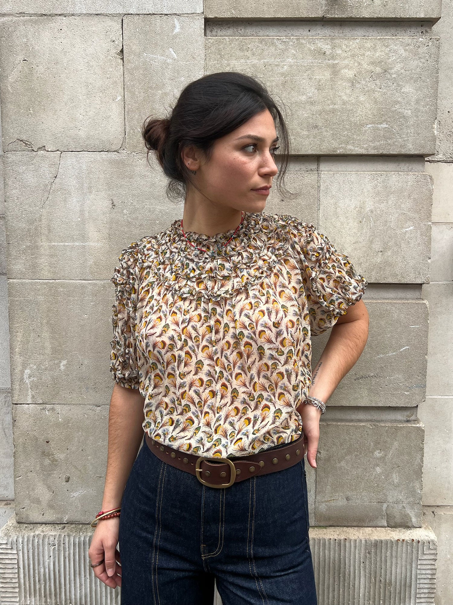 Woman wearing a patterned blouse and jeans standing against a stone wall.