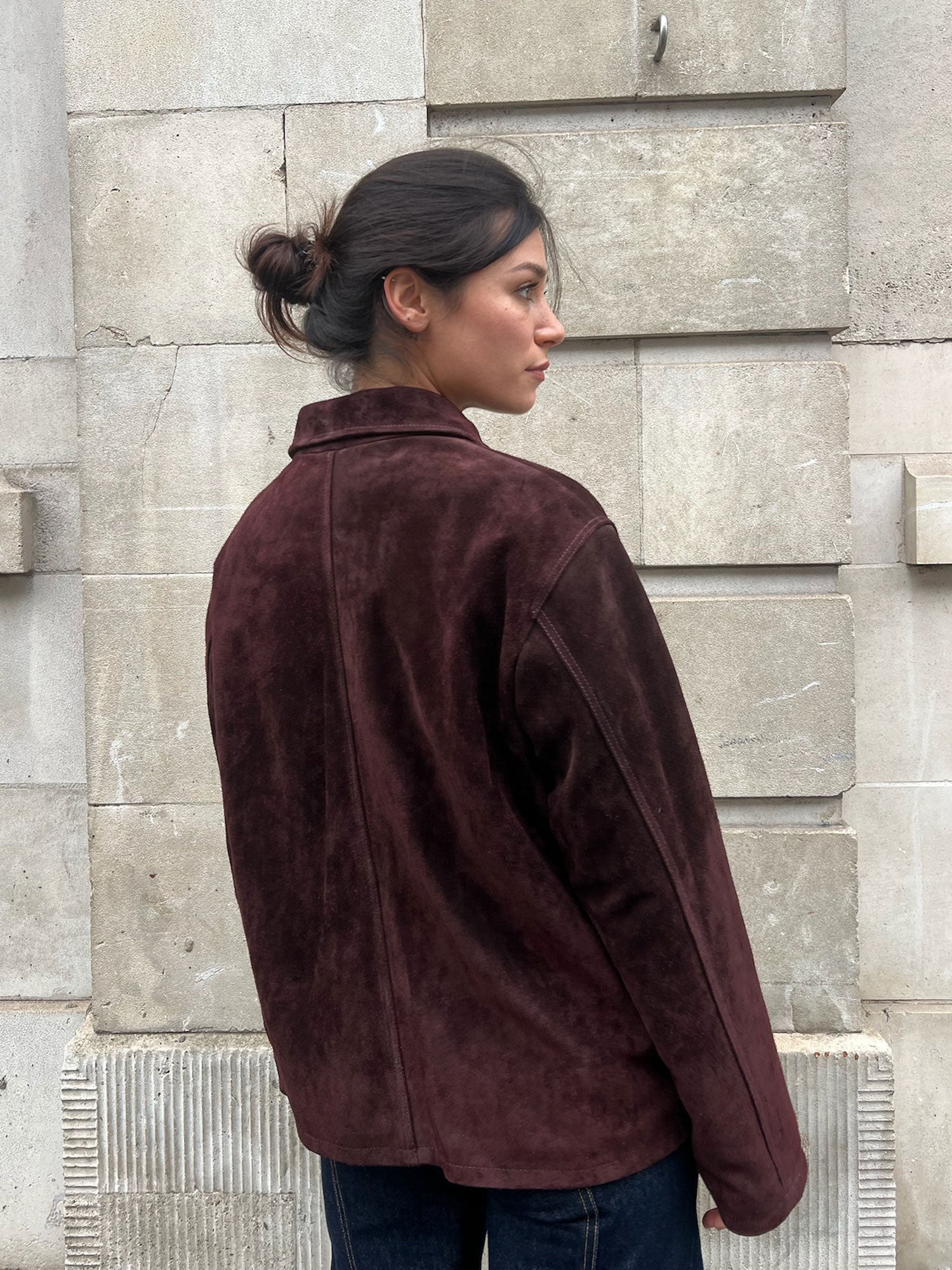 Person wearing a brown jacket standing against a stone wall.