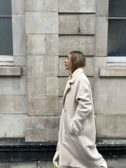 Woman in a beige coat standing against a stone wall.