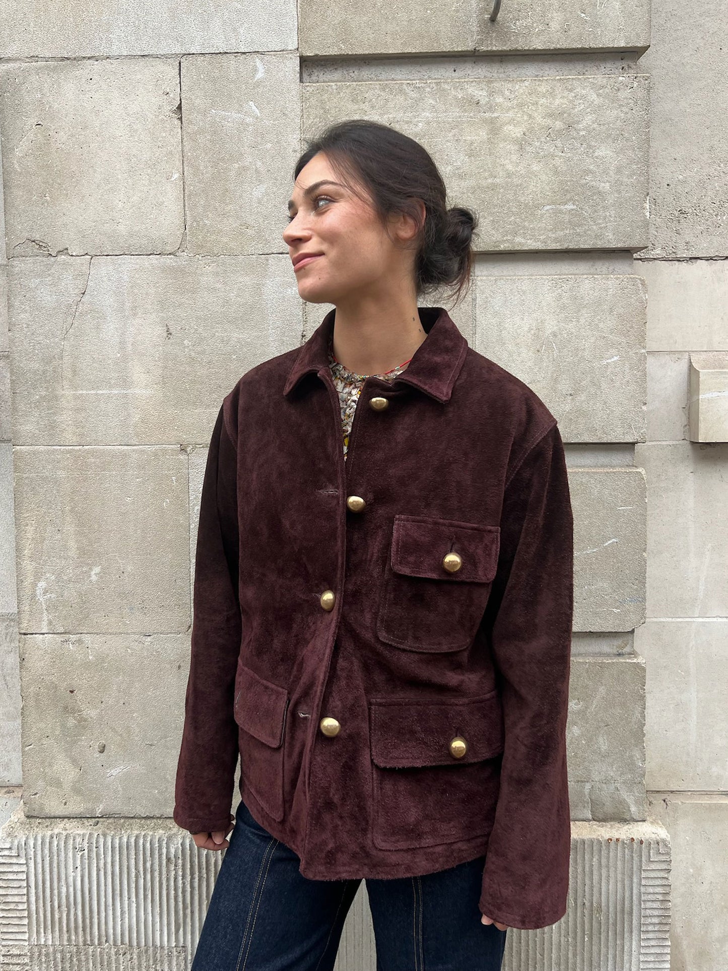 Woman wearing a brown jacket with gold buttons against a stone wall.