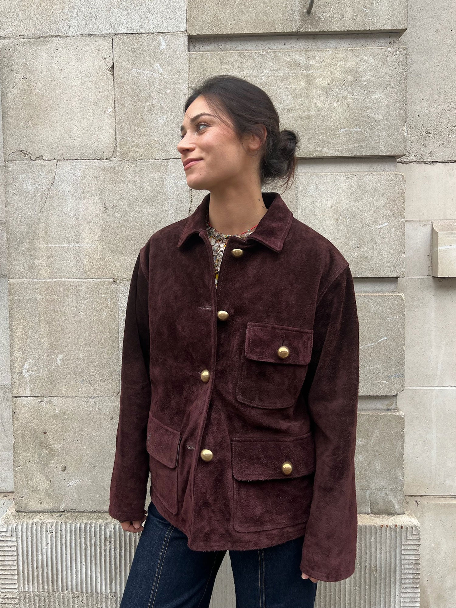 Woman wearing a brown jacket with gold buttons against a stone wall.