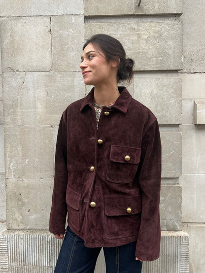 Woman wearing a brown jacket with gold buttons against a stone wall.