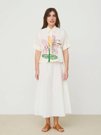 Woman wearing a white Mii shirt  with colorful floral design and a white skirt on a plain background