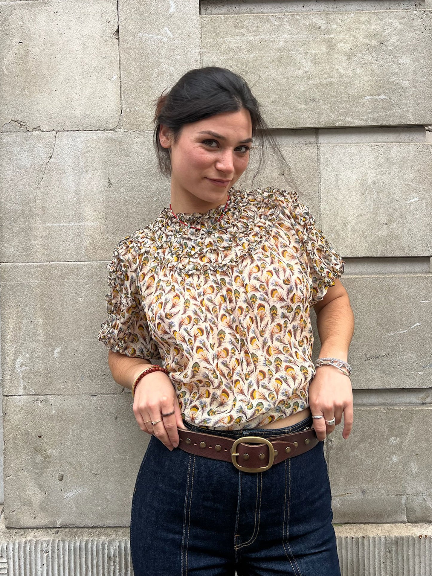 Woman wearing a patterned blouse and jeans standing against a stone wall.
