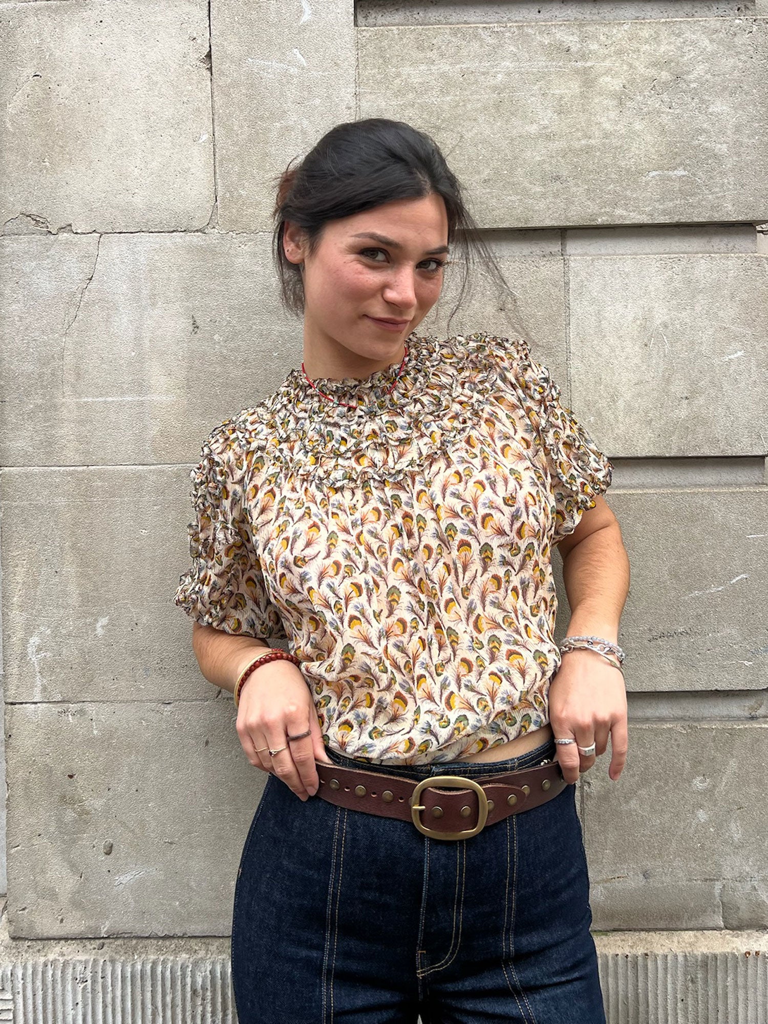 Woman wearing a patterned blouse and jeans standing against a stone wall.